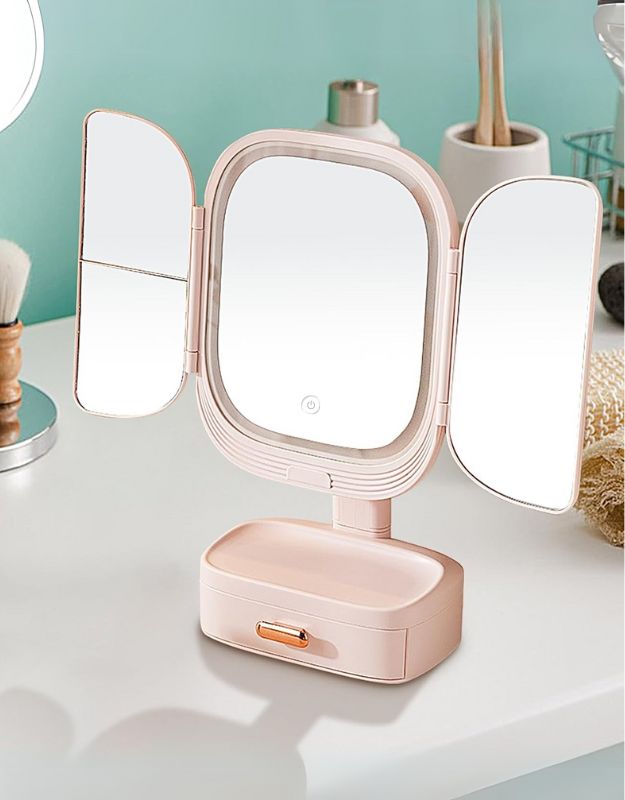 A LED lighted mirror with a foldable design, featuring a small drawer at the base and illuminated panels on either side. The mirror is placed on a countertop with various beauty products around it.